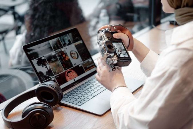 Une photographe tient son appareil photo numérique devant son ordinateur portable pour trier ses clichés.