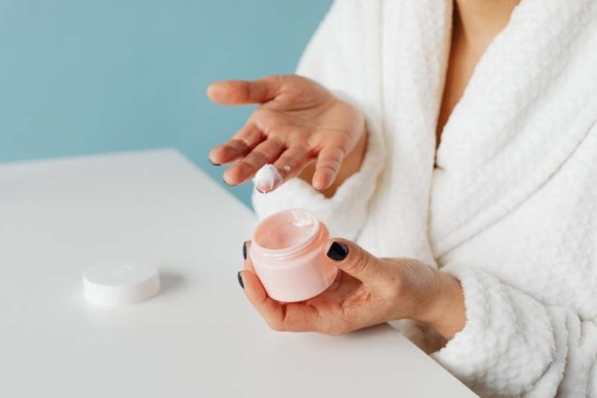 Une personne en peignoir tient un pot de crème hydratante et montre une noisette de produit sur son doigt.