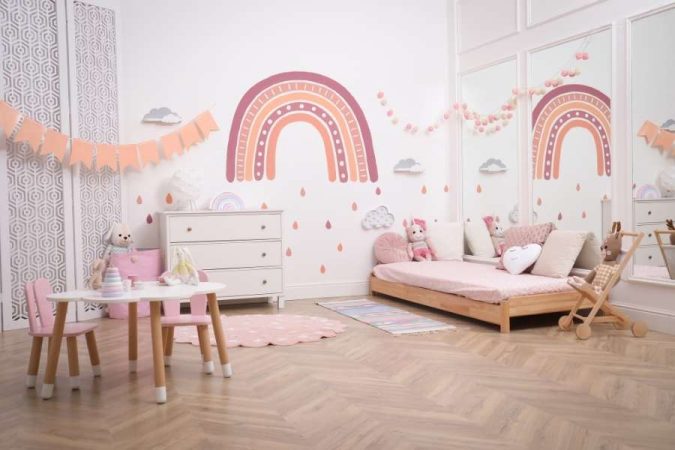 Un lit au sol enfant 90x190 en bois clair avec une literie rose, dans une chambre décorée d'un grand arc-en-ciel mural.