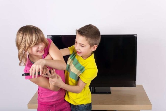Un garçon et une fille se disputent vivement une télécommande devant un téléviseur, illustrant la jalousie entre frères et sœurs.
