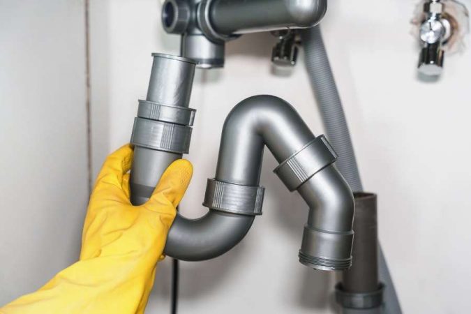 Une main gantée de jaune présente un siphon de lavabo en plastique gris lors de son assemblage.